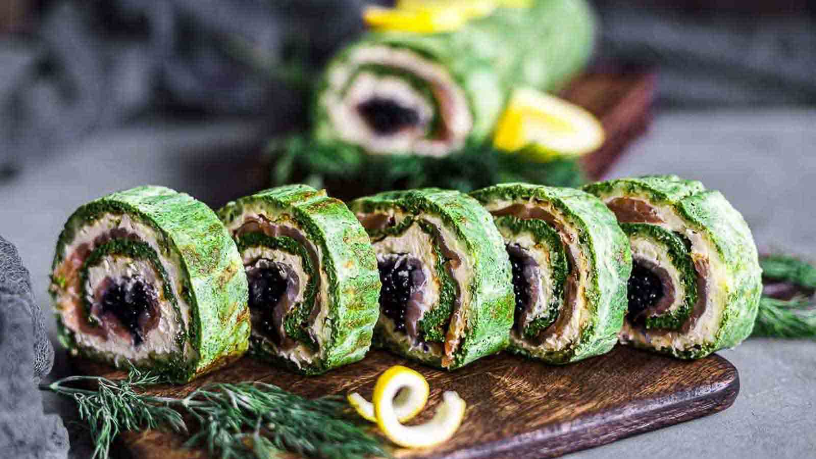 Slices of a rolled green savory dish are neatly arranged on a wooden board, garnished with fresh herbs and lemon slices. The roll appears to be filled with a creamy center and black caviar. Additional uncut roll is visible in the background.
