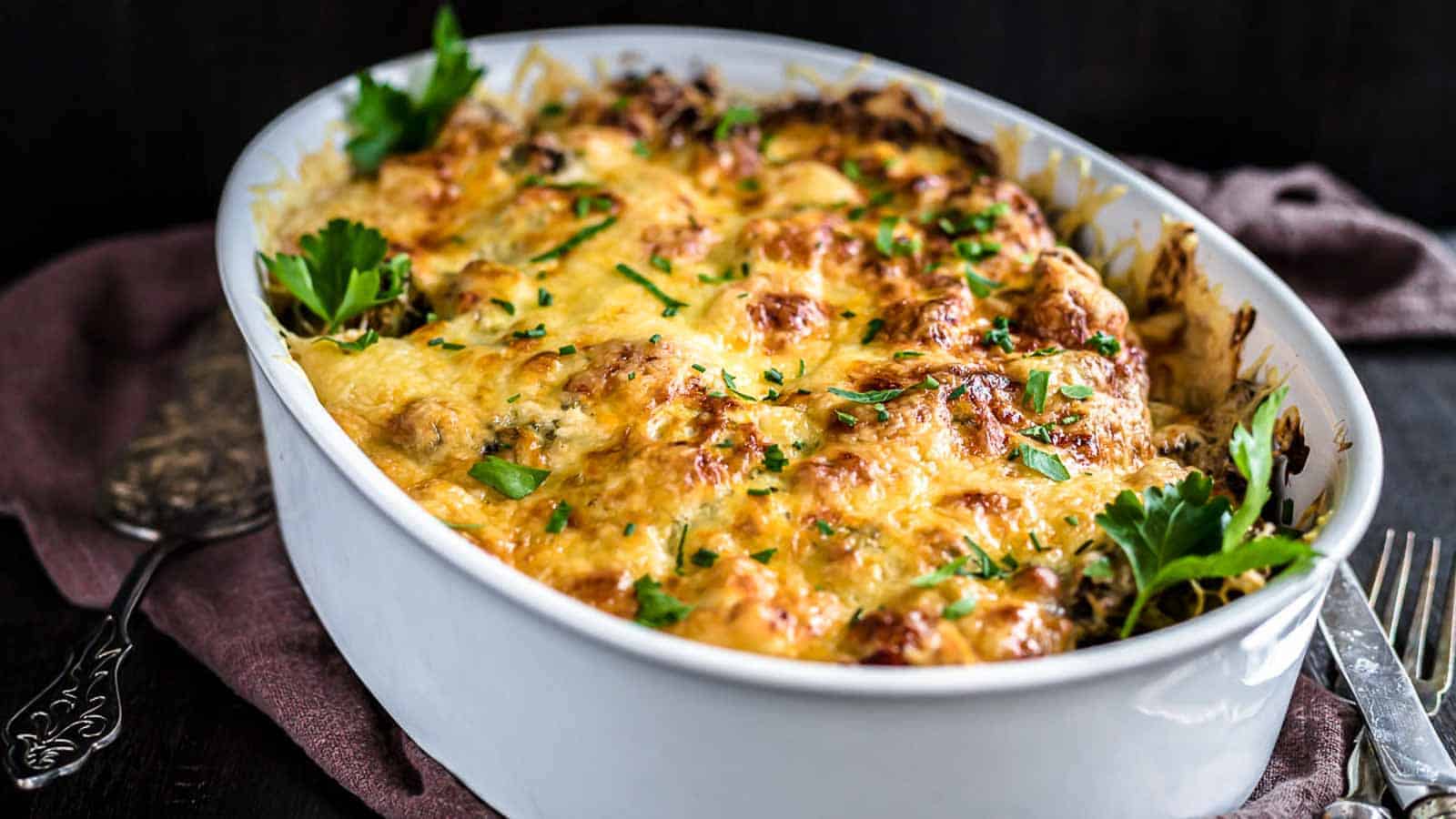A white oval dish is filled with a baked casserole, topped with melted cheese and garnished with fresh parsley. The dish is placed on a dark table with a maroon cloth and silver utensils beside it. The golden-brown, bubbly cheese hints at a delicious meal.