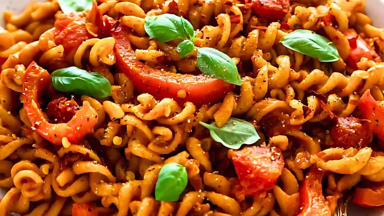 A close-up of a dish featuring cooked spiral pasta mixed with diced tomatoes and garnished with fresh basil leaves. The pasta is coated in a red sauce, giving it a vibrant and appetizing appearance.