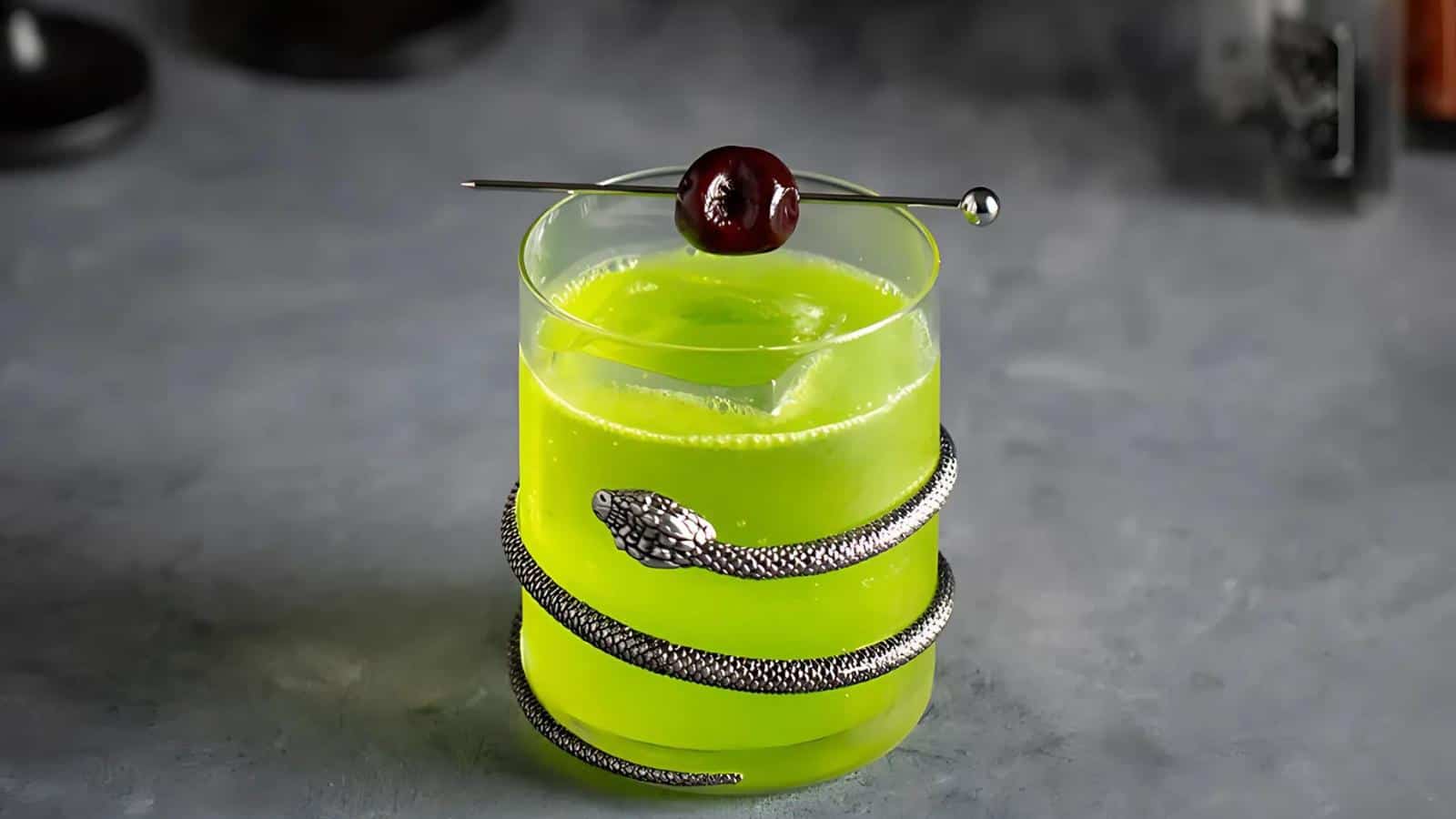 A bright green cocktail in a clear glass is adorned with a silver snake accessory wrapped around the base. A small dark cherry is skewered on a metallic pick resting across the top of the glass. The background is a neutral gray surface.