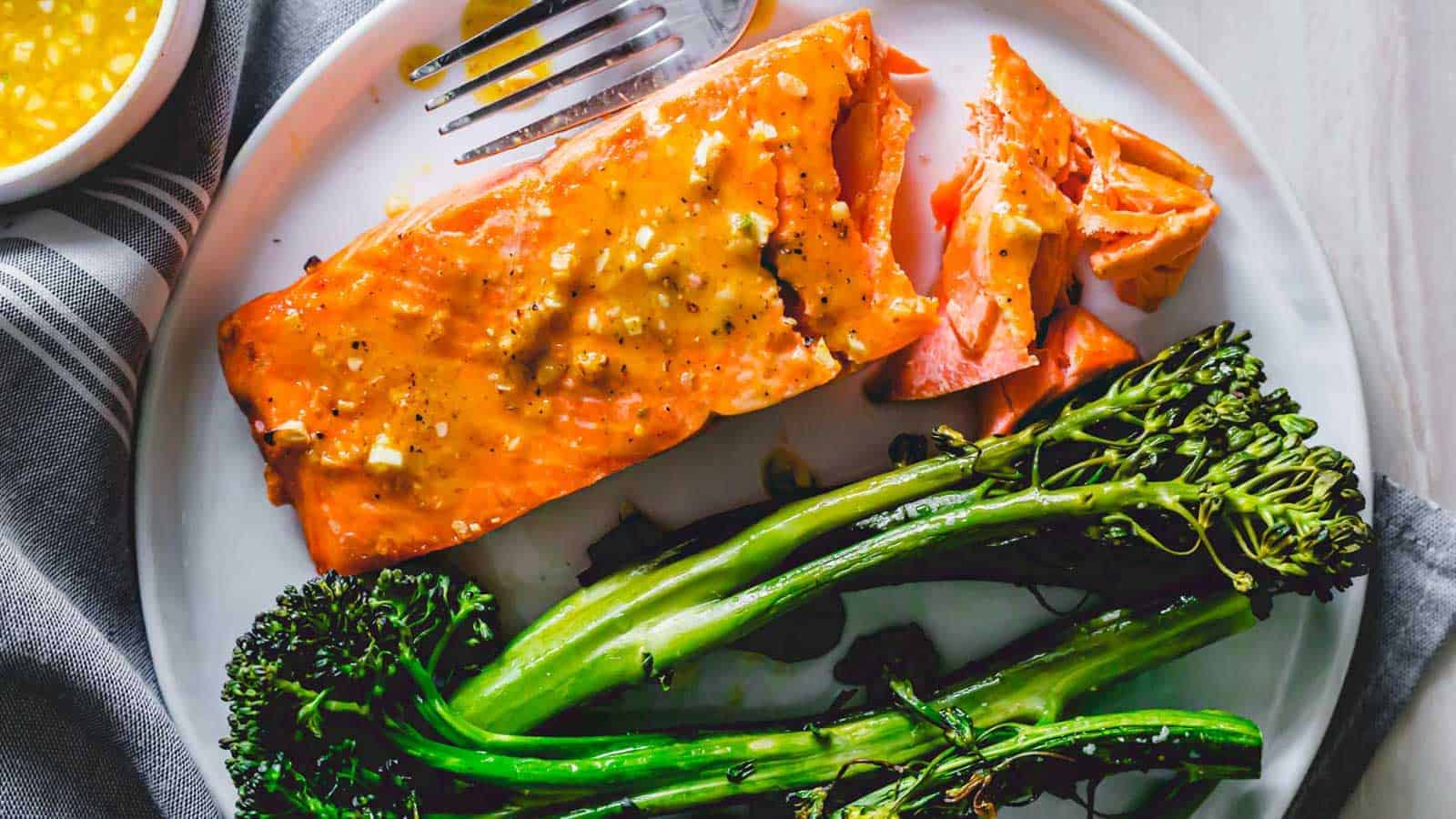 A plate featuring a piece of cooked salmon topped with a mustard-based sauce and seasoned with pepper, accompanied by broccolini. A fork rests above the salmon, and there is a bowl of yellow sauce with a spoon on the side. The plate is on a gray and white striped napkin.