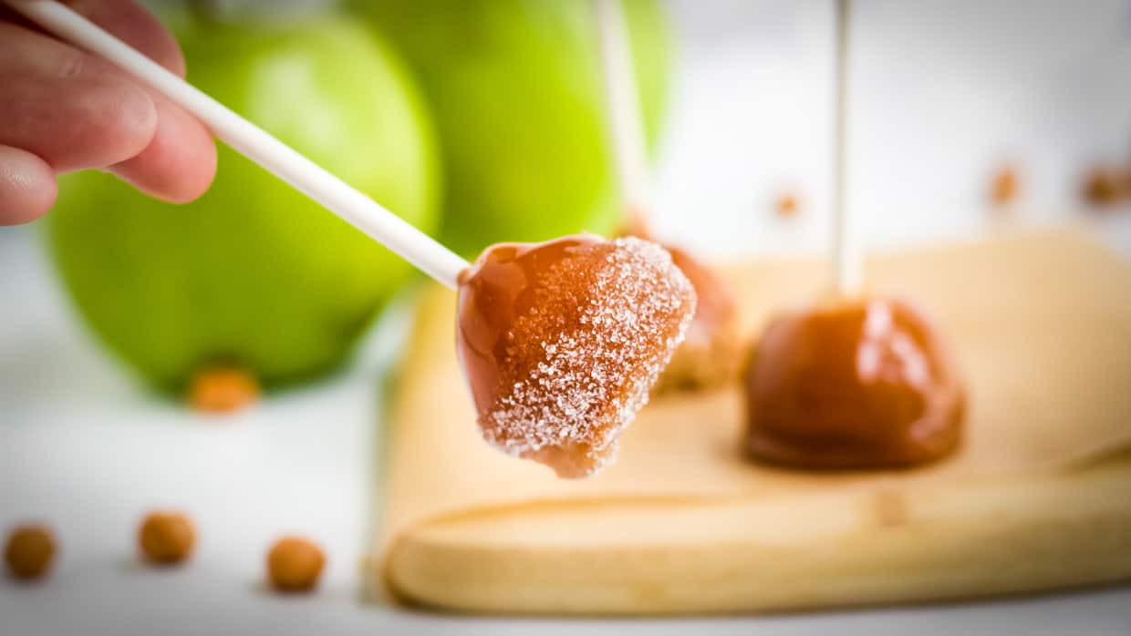 A hand holding a caramel apple pop coated in sugar with more caramel apple pops and green apples blurred in the background. The caramel pops are on white sticks and set on a light wooden surface.