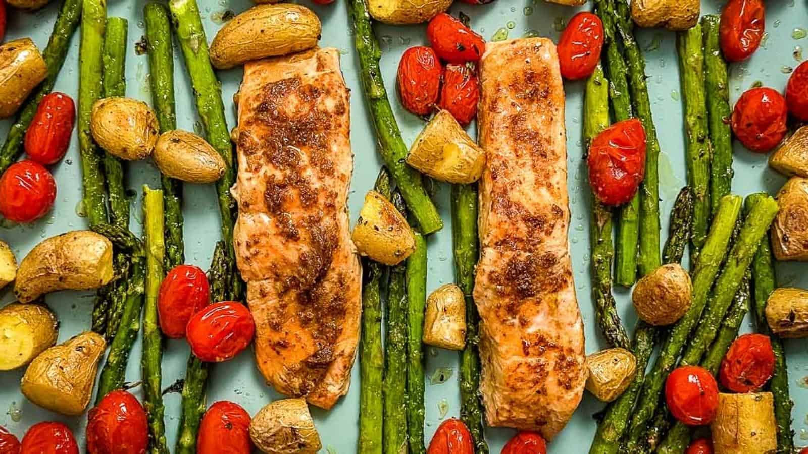 Two seasoned salmon fillets are laid out on a baking sheet with roasted cherry tomatoes, asparagus, and baby potatoes. The dish is vibrant with fresh, colorful vegetables surrounding the salmon, showcasing a healthy, oven-roasted meal.