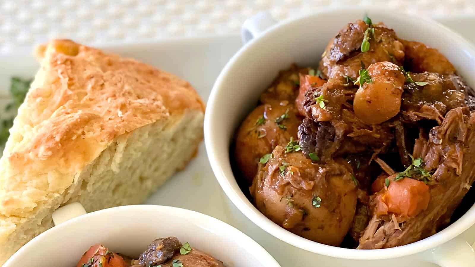 A close-up of a richly colored beef stew with potatoes and carrots served in a white bowl. A slice of golden brown bread is placed on the side. The dish is garnished with chopped herbs.