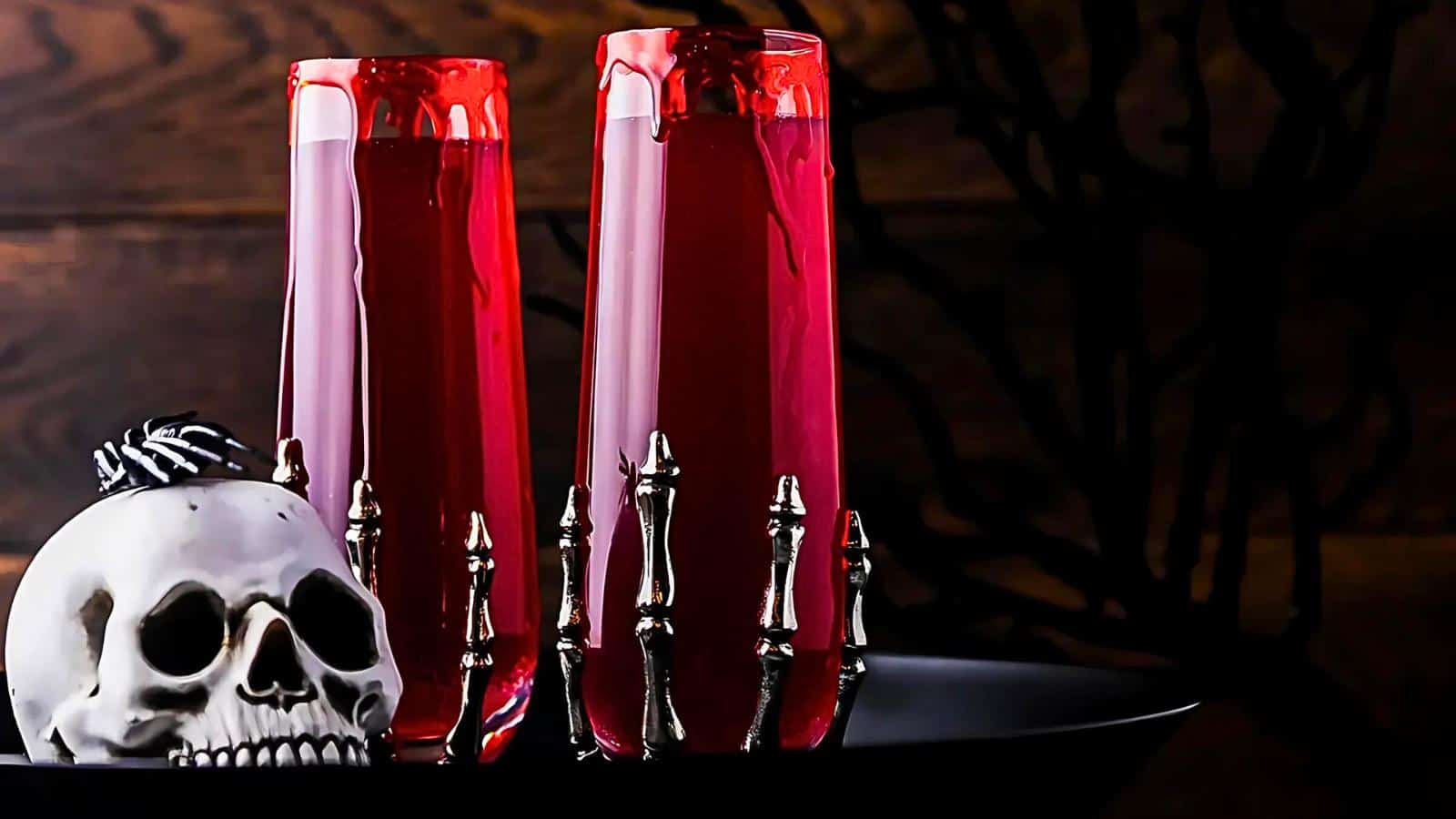 Two tall glasses filled with red liquid stand on a black tray, adorned with skeletal hands clutching the glasses. Beside them, a white skull with a small black decoration on top adds a spooky Halloween vibe. The background is dark and eerie.