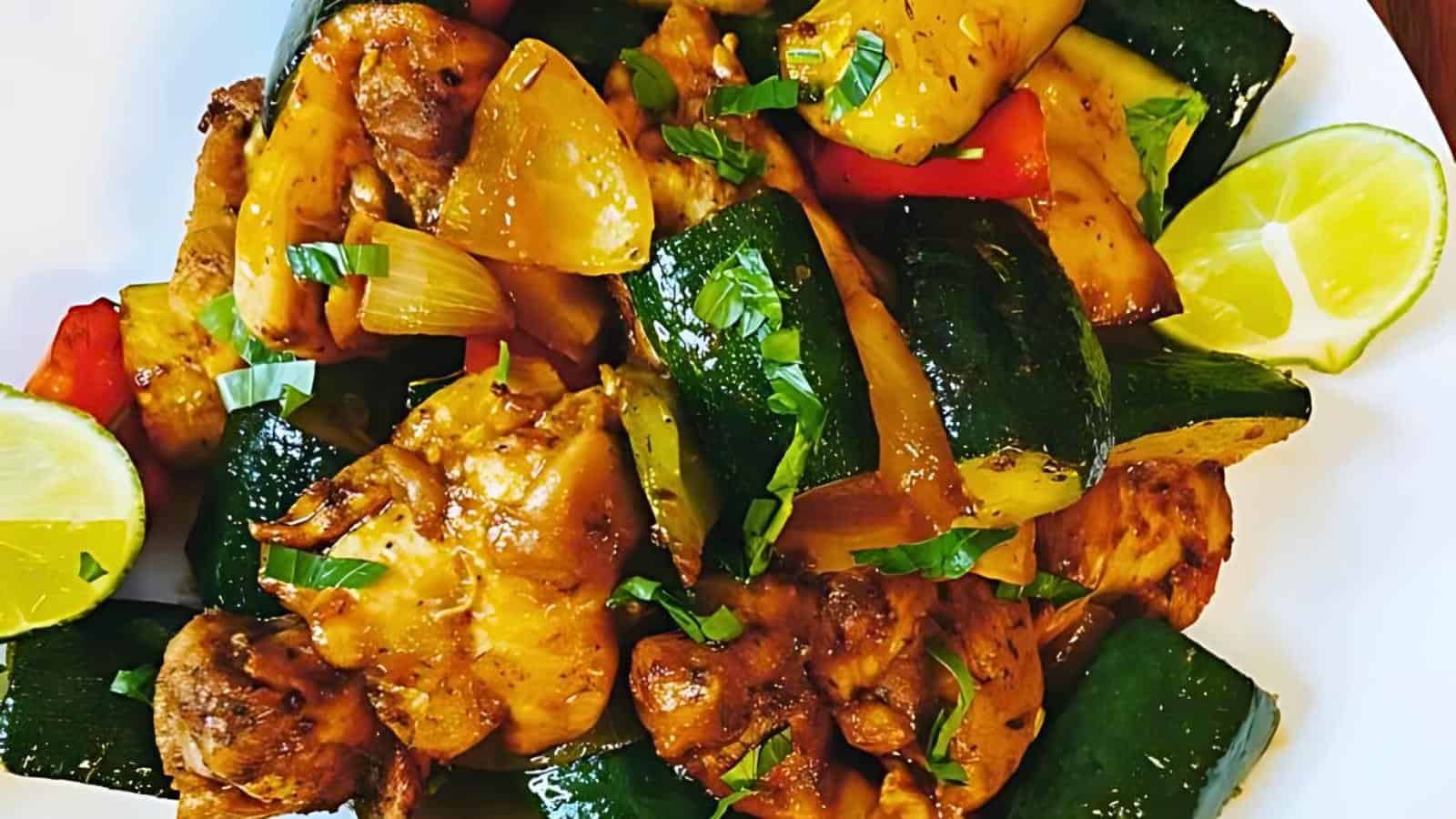 A vibrant dish featuring marinated chicken chunks and assorted vegetables, including zucchini, bell peppers, and onions, all garnished with fresh herbs. Lime wedges are provided on the side for an added citrus flavor. The dish is presented on a white plate.