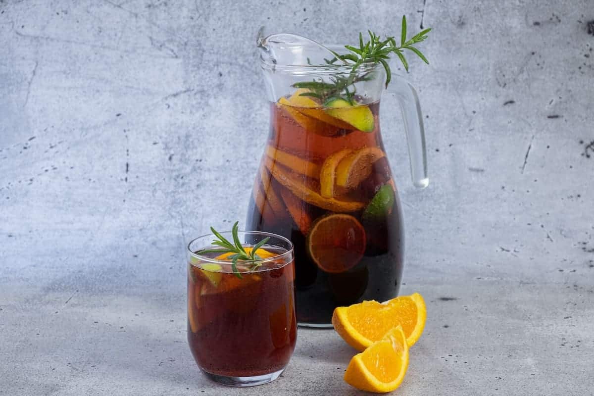 A glass pitcher and a small glass filled with a dark iced beverage, garnished with slices of orange, lemon, and a sprig of rosemary. Orange wedges and rosemary are placed next to the glass on a grey, textured surface.