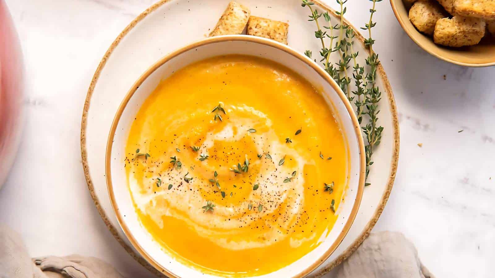 A bowl of creamy pumpkin soup garnished with black pepper and fresh thyme. The bowl is placed on a white plate with a side of croutons on the plate. Another bowl of croutons, a napkin, and a hint of a pink object are visible in the background.