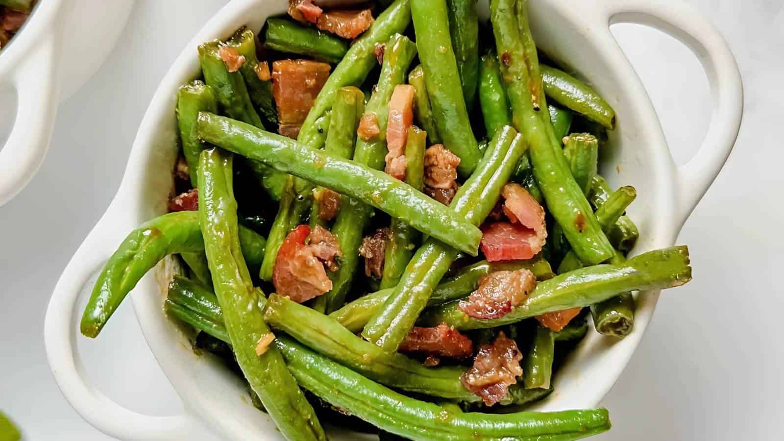 A white bowl filled with cooked green beans mixed with chunks of crispy bacon. The green beans have a glossy appearance, indicating they might be sautéed, and the bacon pieces add texture and a pop of color to the dish.