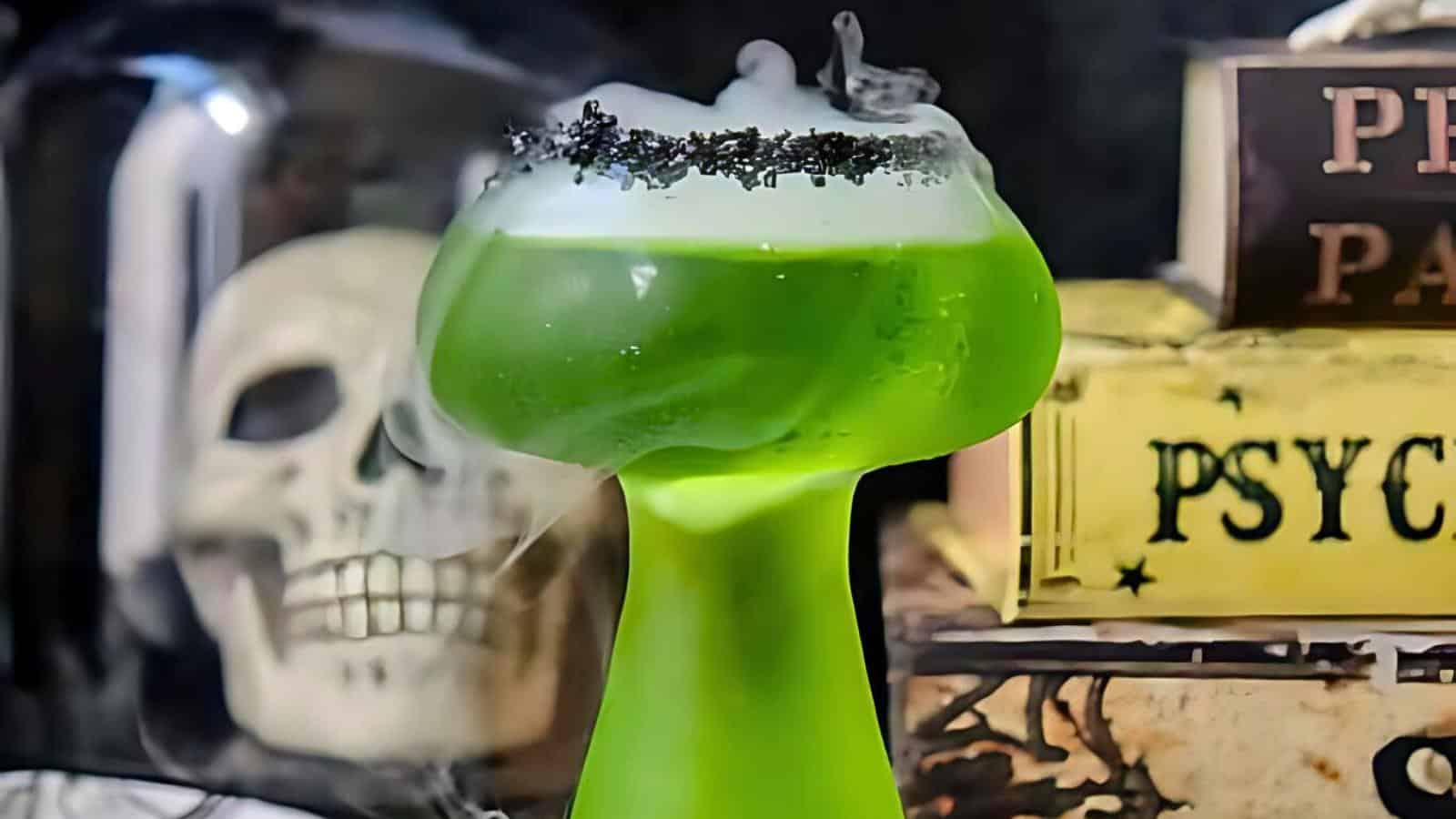 A green, foamy drink served in a goblet with a black rim sits in front of a spooky background featuring a skull and vintage books, one of which has "PSYCH" in bold letters. Atmospheric smoke adds an eerie effect to the scene.