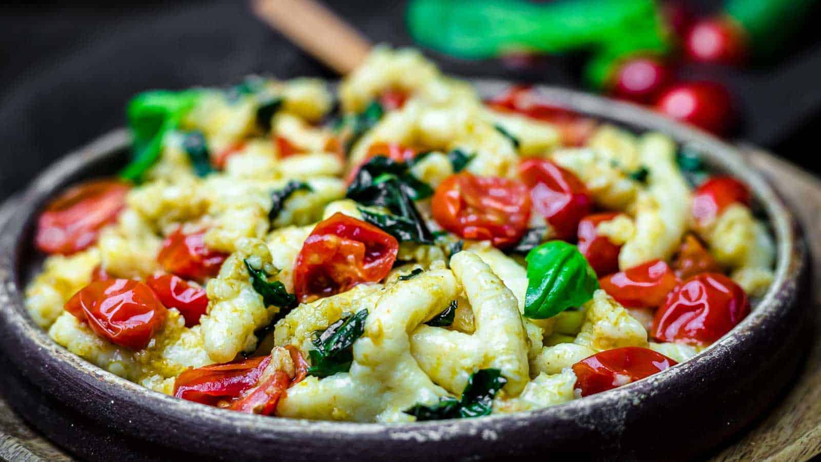 A bowl filled with gnocchi mixed with vibrant cherry tomatoes and fresh basil leaves, all delicately cooked, is shown in the foreground. The background is slightly blurred but hints at more fresh ingredients, adding depth to the image.