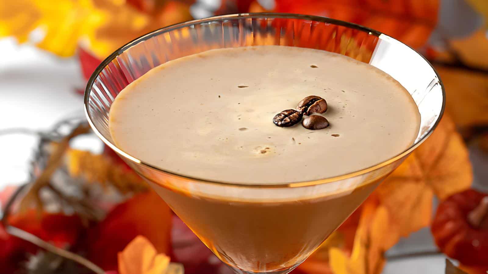 A creamy coffee cocktail served in a martini glass with three coffee beans as garnish. The background features soft focus autumn leaves in warm tones, enhancing the seasonal atmosphere.