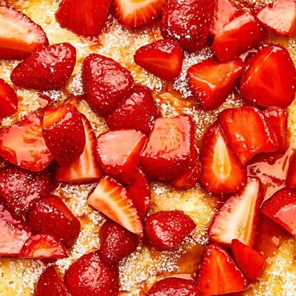 A close-up view of a dessert featuring a layer of sliced strawberries on a golden, slightly caramelized surface. Powdered sugar is dusted lightly over the strawberries and the dessert's crust, creating a sweet and appetizing presentation.