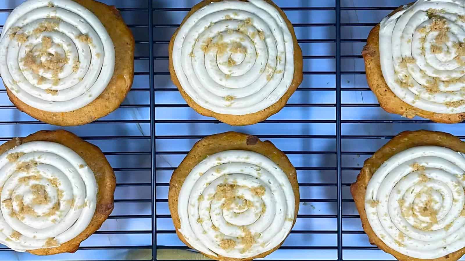 Six freshly baked cookies with swirled icing and sprinkled crumbs on top are cooling on a wire rack. The cookies are evenly spaced in two rows of three, and the wire rack is placed over a blue and white surface.