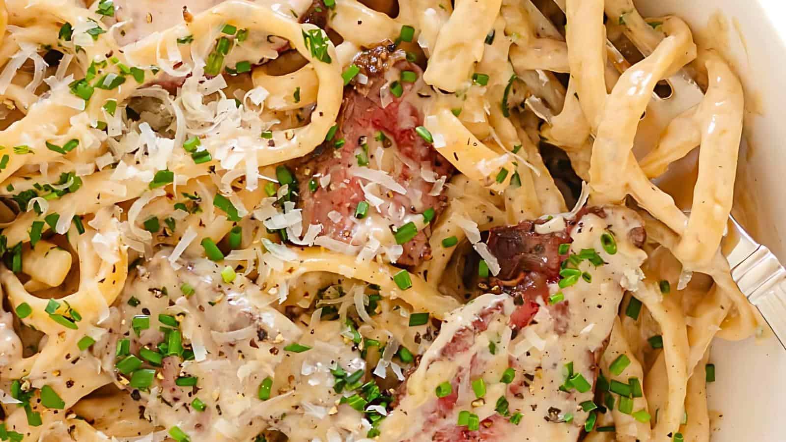 A close-up of a creamy pasta dish garnished with chopped chives. The pasta strands are intertwined with pieces of cooked meat and covered in a rich, white sauce with grated cheese on top. A fork is partially visible to the side, ready to dig in.