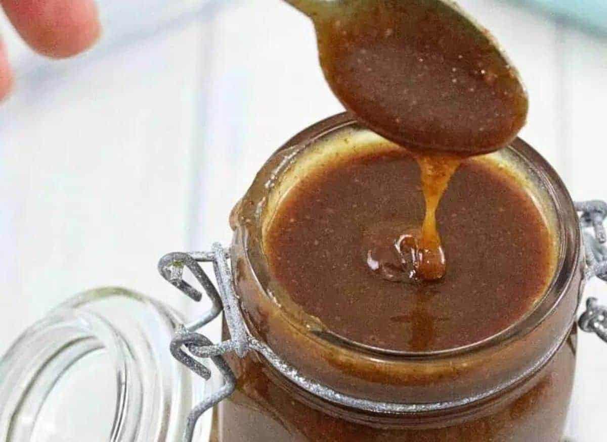 A close-up image of a jar filled with thick, dark brown sauce or syrup. A spoon hovers above the jar, with some of the sauce dripping off it. The jar is secured with a metal clasp. The background is softly blurred.