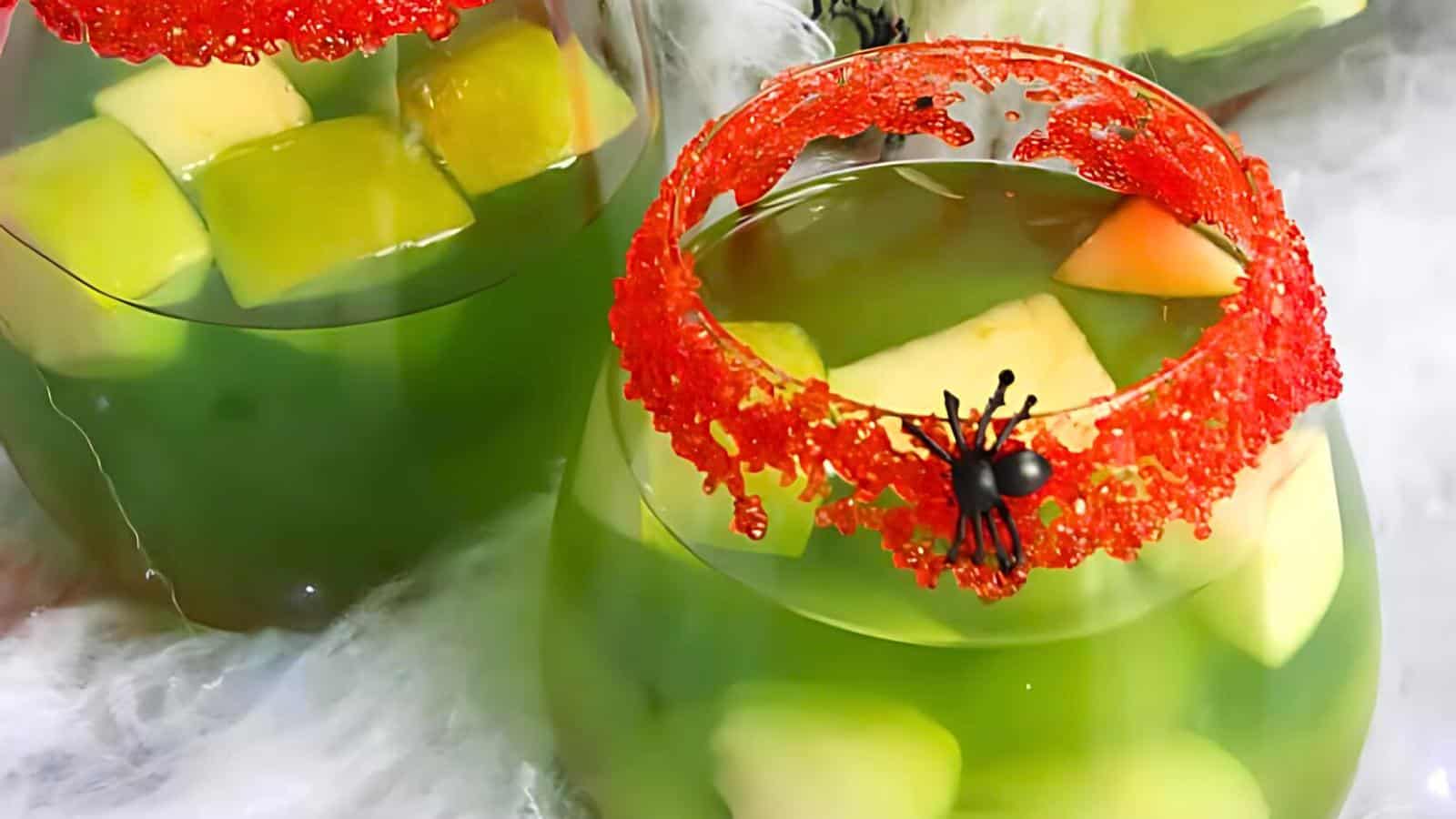 Two glass cups filled with a green liquid and chunks of fruit. The rims of the cups are covered in red sugar crystals, and a small plastic black spider decorates one of the rims. The background includes a white, web-like texture.