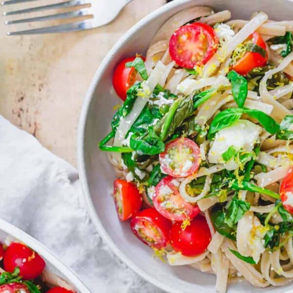 A bowl of linguine pasta topped with cherry tomatoes, fresh basil leaves, and grated cheese. A fork and additional basil leaves are placed next to the bowl on a light-colored wooden surface. The dish is vibrant and garnished with finely chopped herbs.