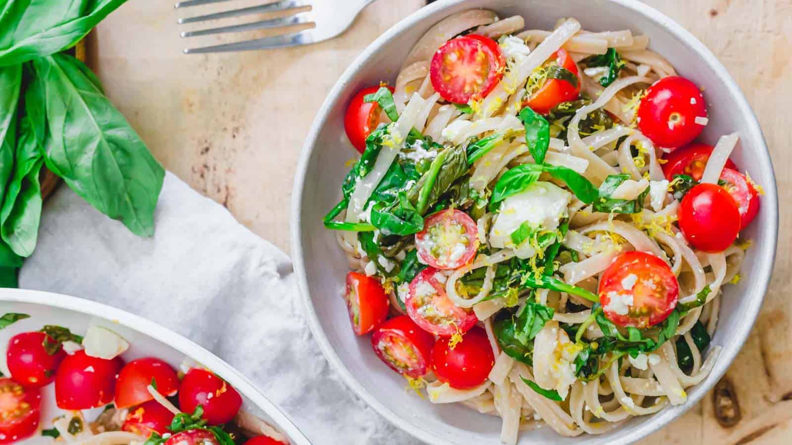 A bowl of linguine pasta topped with cherry tomatoes, fresh basil leaves, and grated cheese. A fork and additional basil leaves are placed next to the bowl on a light-colored wooden surface. The dish is vibrant and garnished with finely chopped herbs.