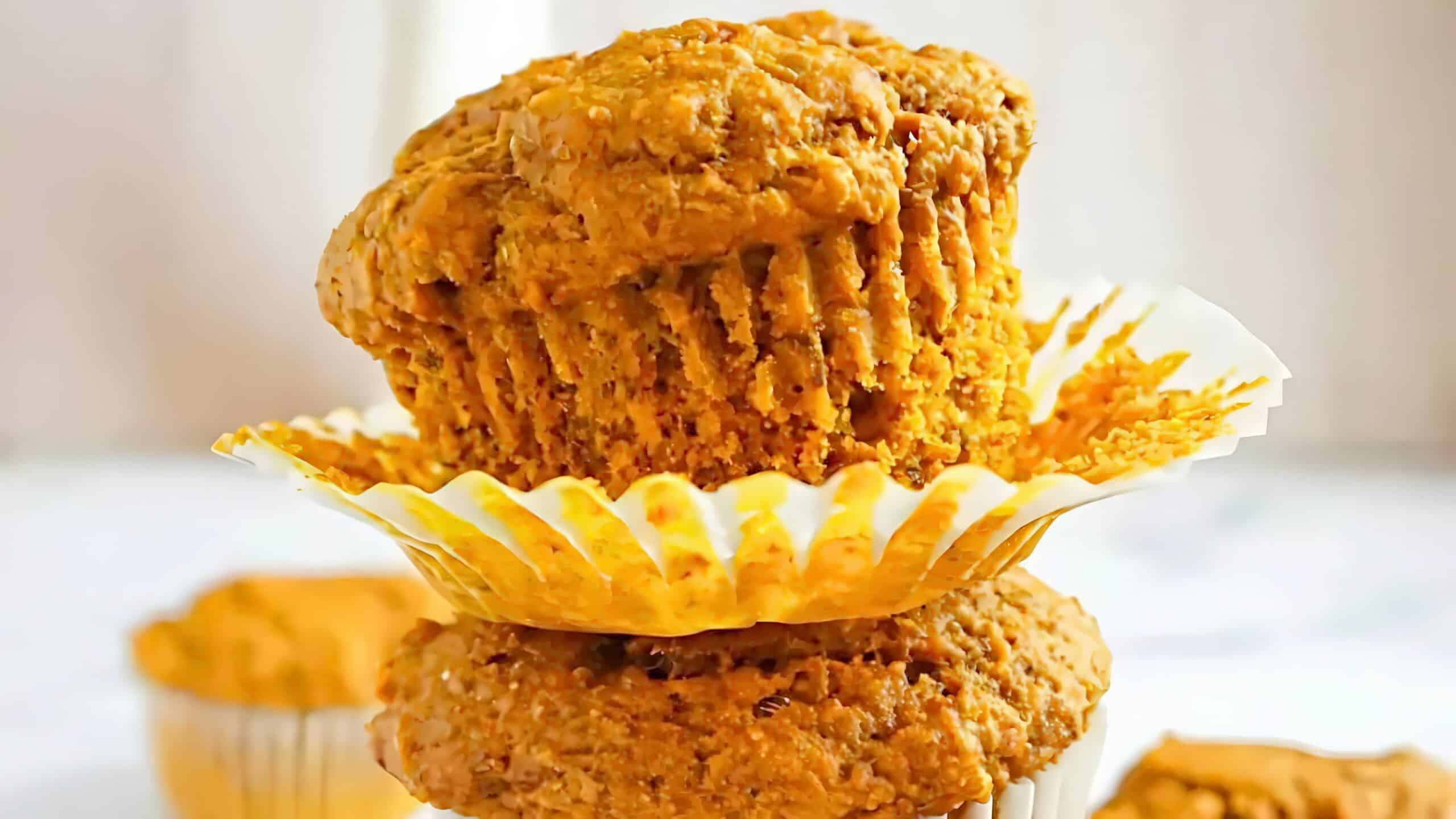 A close-up of two stacked muffins with a light orange hue, each partially wrapped in white paper liners. The top muffin has its liner partially peeled back, showing a moist, textured surface. Another muffin is slightly visible in the background.