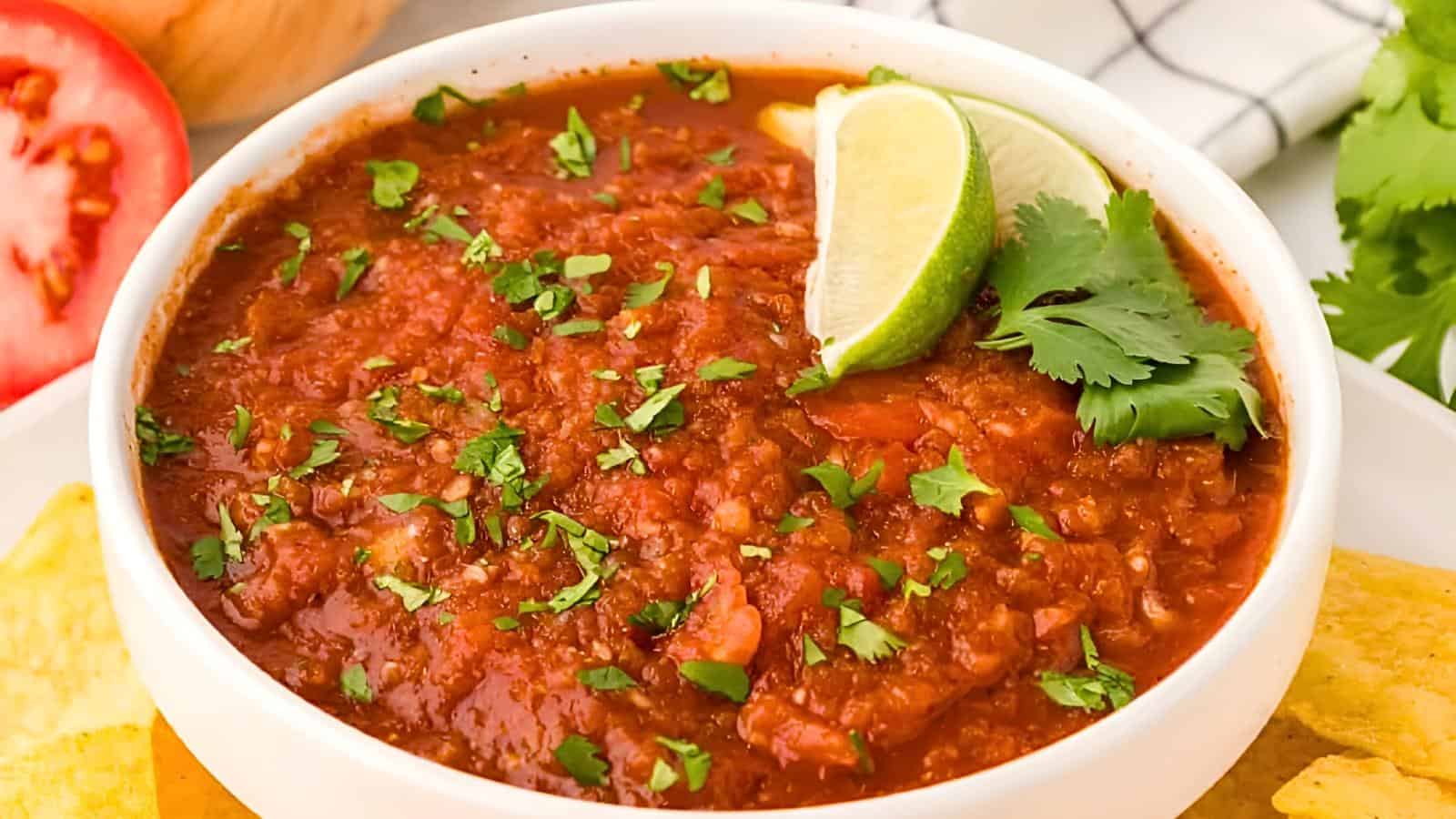 A bowl of vibrant red salsa garnished with chopped cilantro and two lime wedges. The salsa is accompanied by yellow tortilla chips on the side. In the background, a halved tomato, an onion, and cilantro garnish can be seen.