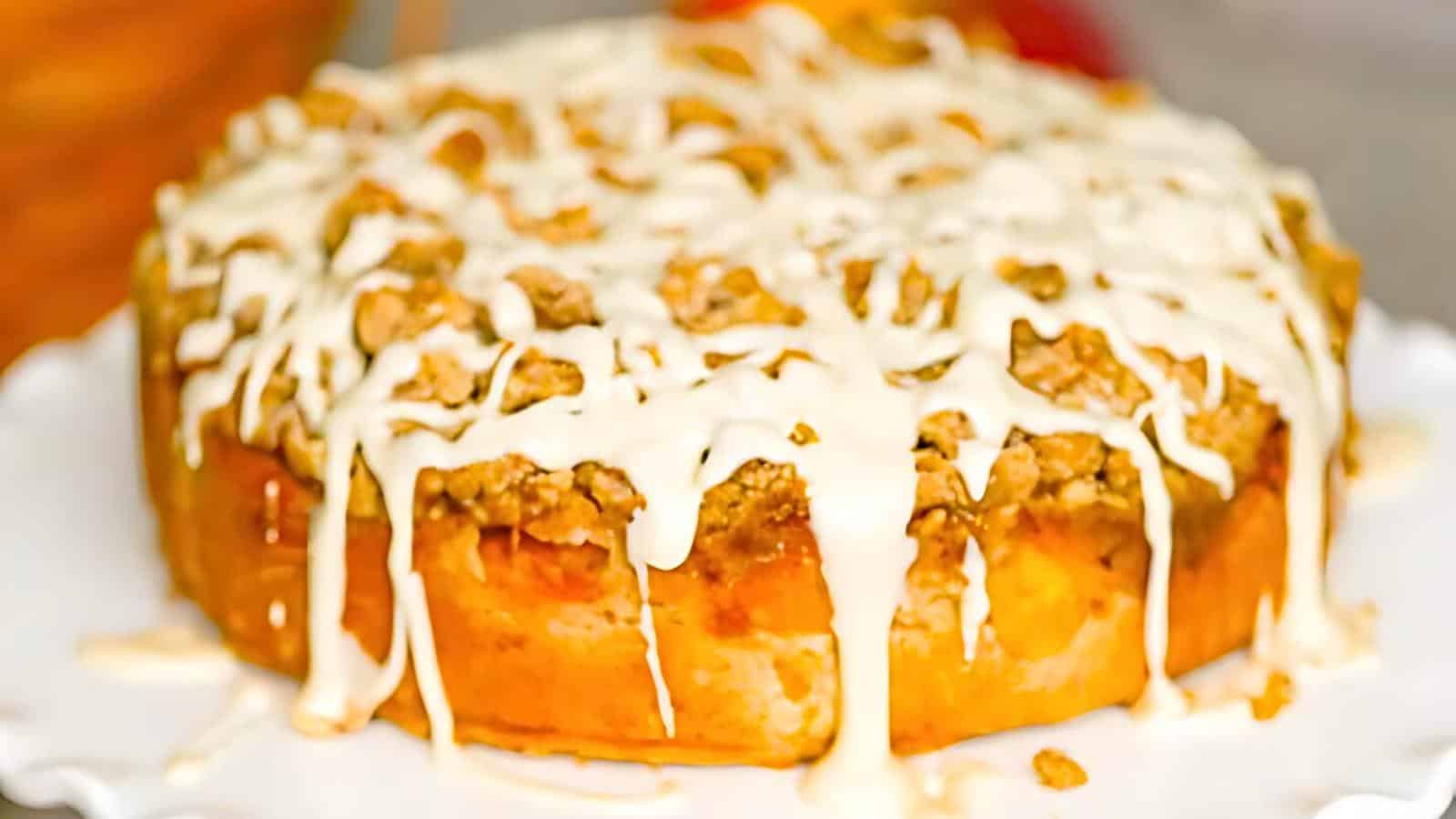 A round, iced coffee cake with a crumbly topping, displayed on a white plate. The icing is drizzled generously over the top, creating a visually enticing dessert.