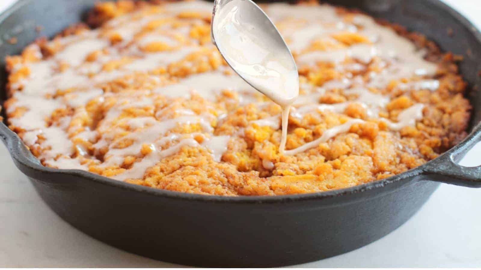 A black cast iron skillet filled with a freshly baked peach cobbler, topped with a drizzle of icing. A spoon held above the skillet is shown mid-drip, adding more icing to the cobbler. The dish has a golden-brown, crumbly texture with visible peach pieces.