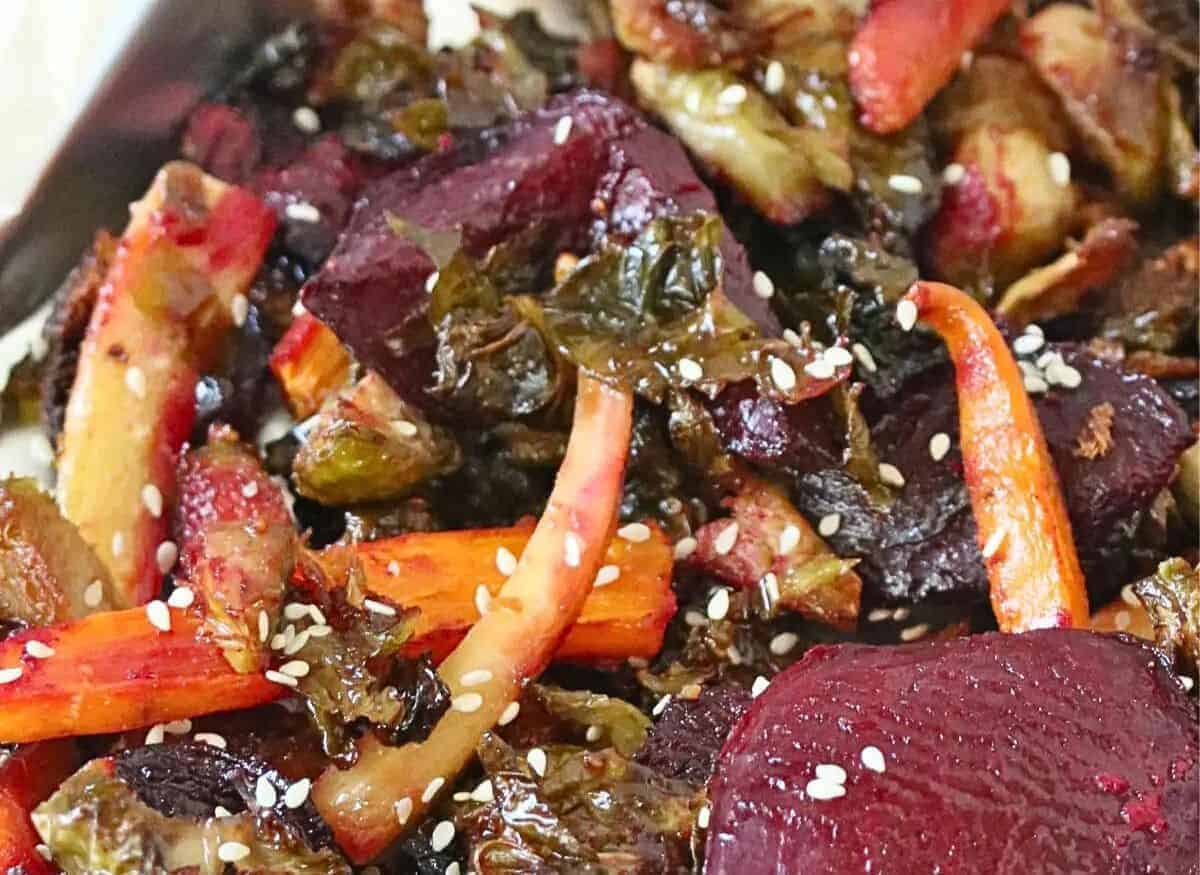 Close-up of a mixed vegetable dish featuring roasted beets, carrots, and leafy greens, garnished with sesame seeds. The vegetables are caramelized and appear glazed, showcasing rich colors and textures.