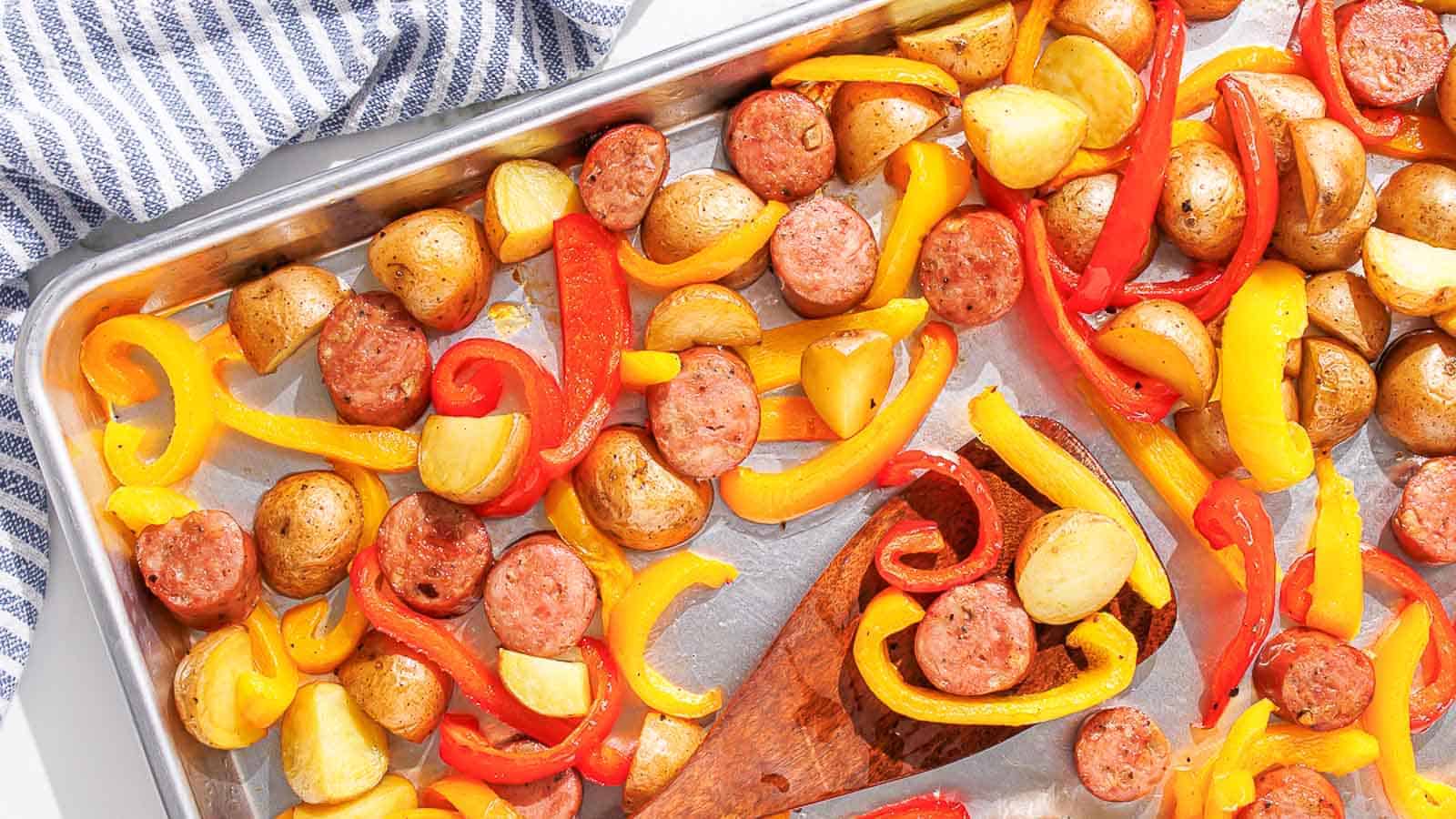 A baking sheet filled with roasted baby potatoes, slices of sausage, and strips of red and yellow bell peppers, all seasoned and cooked to a golden brown. A wooden spatula is also on the sheet, with a blue and white striped cloth visible in the corner.