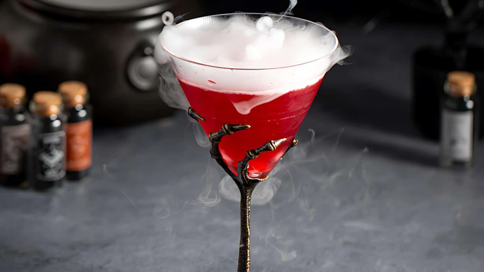 A spooky cocktail in a glass with a skeletal hand as the stem, filled with a red liquid and topped with a layer of smoke-like fog. The background features small bottles, adding to the eerie atmosphere.