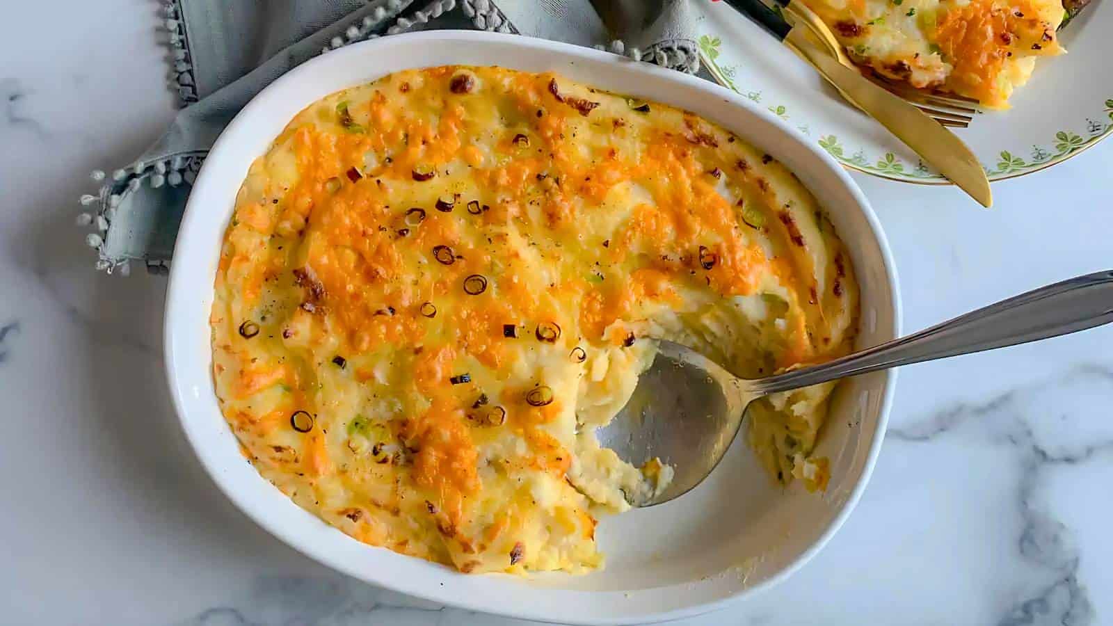 A white baking dish filled with mashed potatoes topped with melted cheddar cheese and garnished with chopped green onions. A silver serving spoon rests on the right side of the dish, where a portion has been scooped out. A portion is served on a nearby plate.