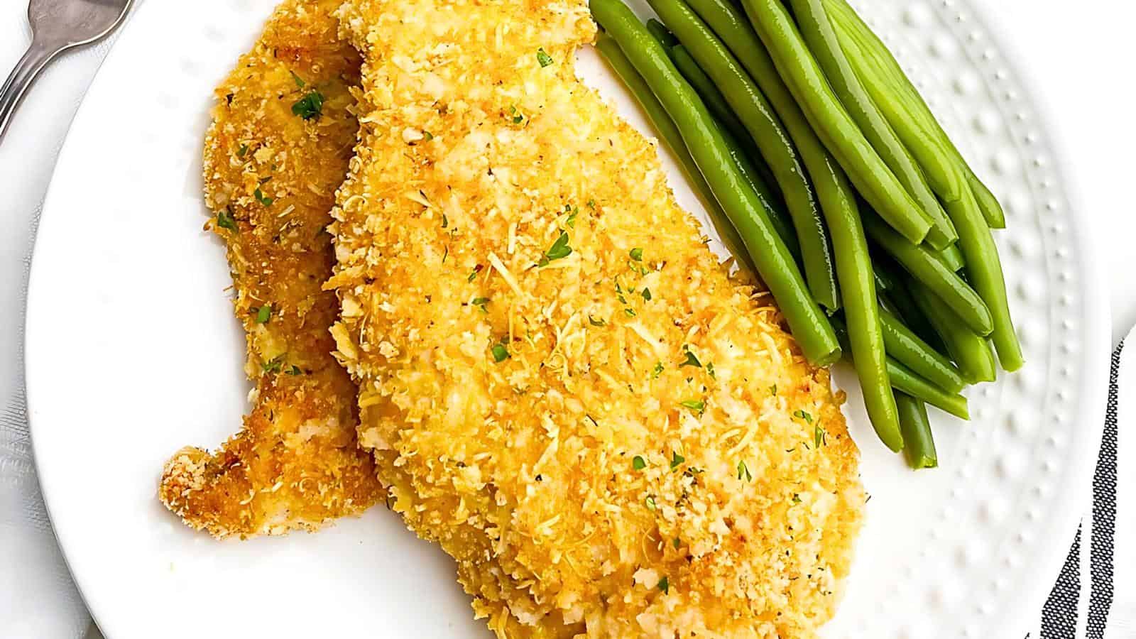 A white plate holds two pieces of golden-brown Parmesan-crusted chicken and a serving of green beans. The chicken is topped with a light sprinkling of herbs. The edge of a fork and a striped napkin are partially visible in the background.
