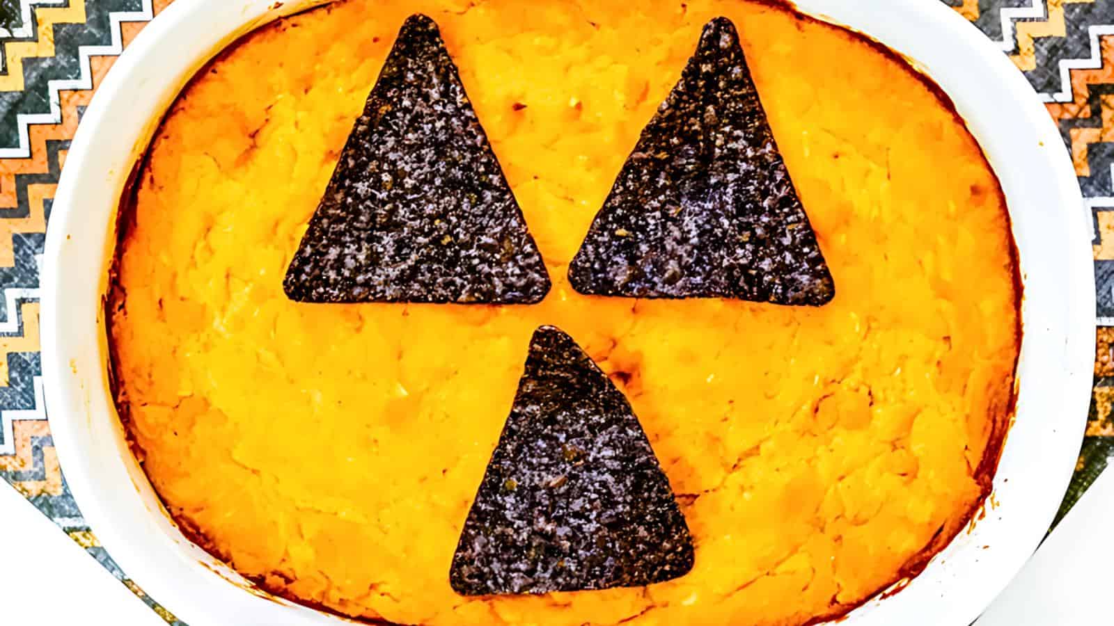 A casserole dish filled with a bright orange mash, possibly sweet potatoes, topped with three dark, triangular tortilla chips arranged to resemble a jack-o'-lantern face. The dish sits on a patterned, zigzag tablecloth.