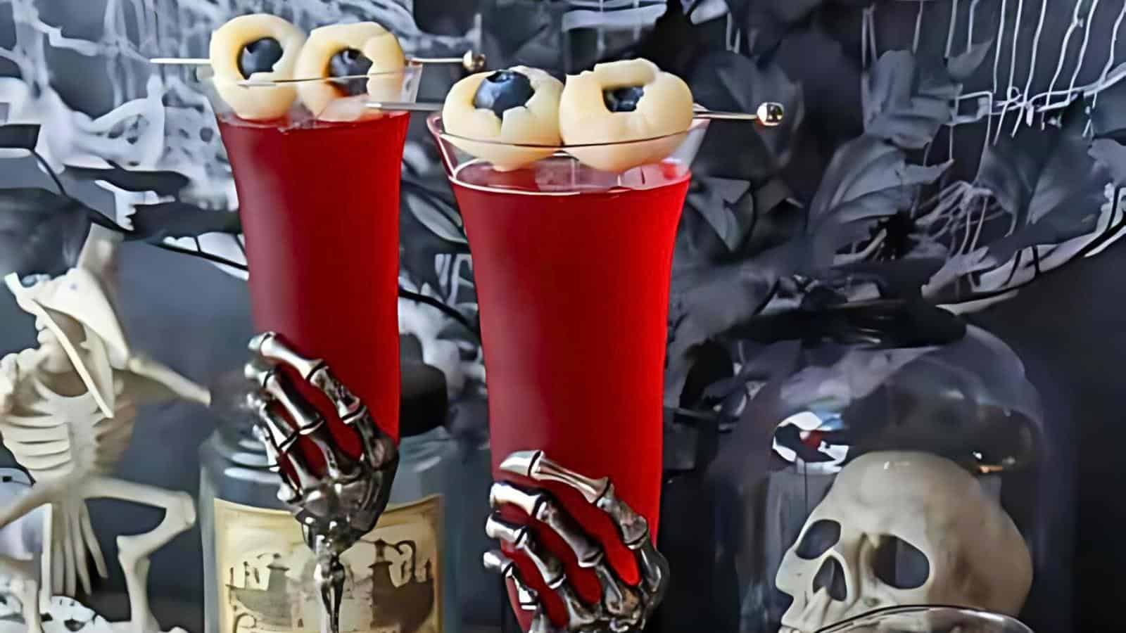 Two tall glasses filled with red liquid and garnished with skewered lychee eyeballs. The glasses are held by skeletal hands. The background includes a spooky skull decoration, a skeleton figure, and dark, eerie decor.