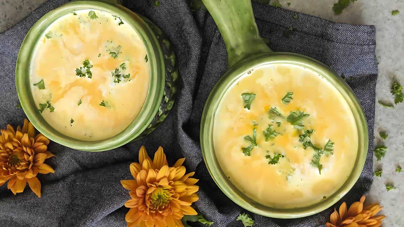 Two bowls of creamy soup garnished with fresh herbs in green ceramic bowls are placed on a dark cloth. Bright orange flowers are artistically arranged around the bowls, creating a warm and inviting presentation. The background appears to be a gray surface.