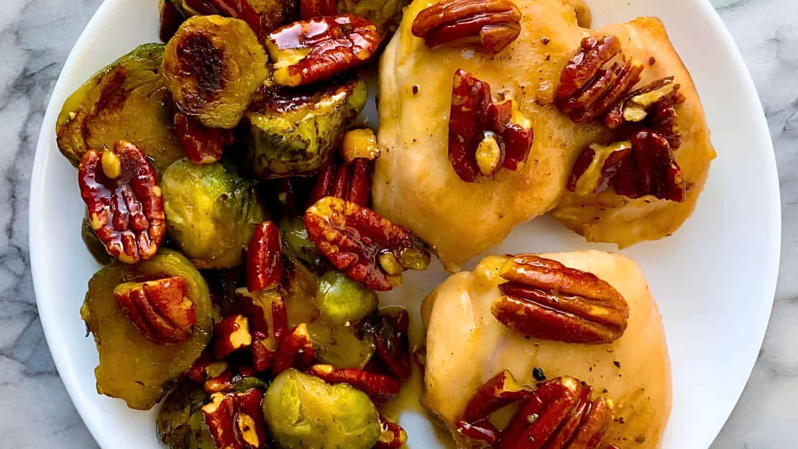 A plate of roasted Brussels sprouts and chicken thighs, topped with a mixture of caramelized pecans. The sprouts are browned and the chicken is cooked to a golden color. The dish is garnished with the glossy pecans, adding a rich glaze to the meal.