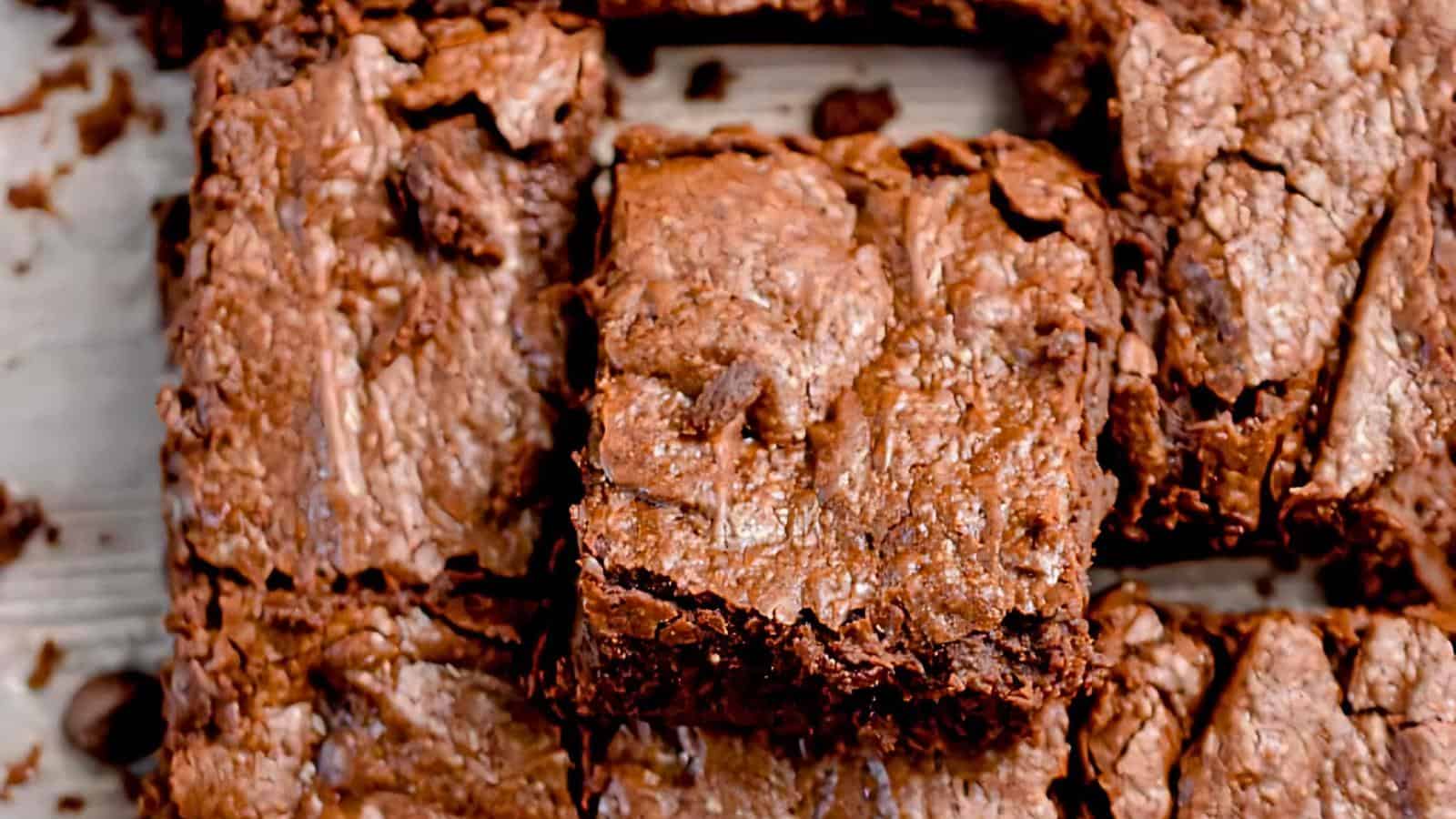 Close-up of freshly baked, fudgy chocolate brownies cut into squares on a wooden surface. The brownies have a rich, glossy top and a slightly cracked texture, highlighting their gooey, dense interior. Crumbles of brownies are scattered around.