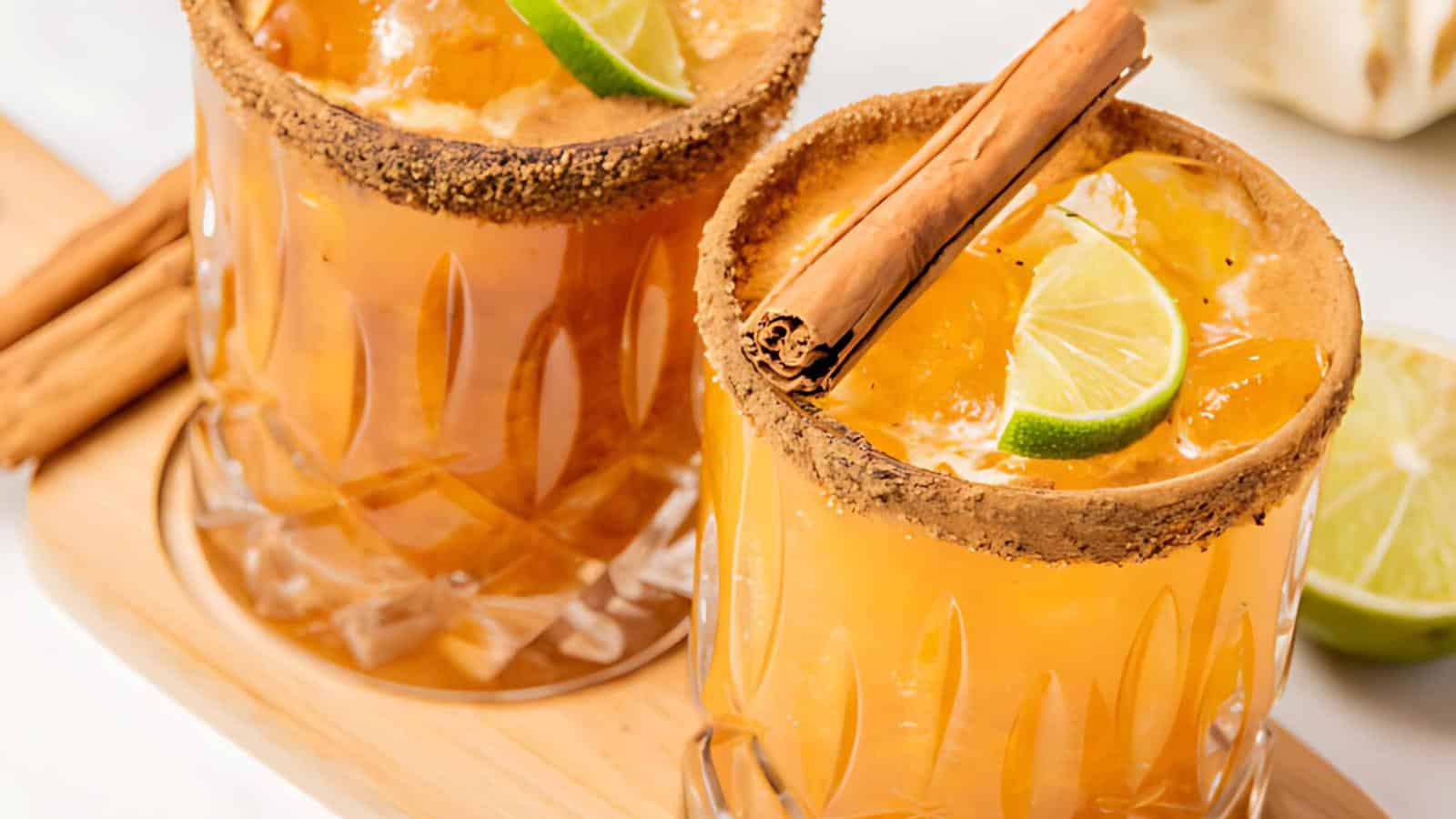 Two glasses of iced beverage, garnished with lime wedges and cinnamon sticks, sit on a wooden tray. The glasses have a brown sugar rim and are filled with an orange-colored liquid. There are additional cinnamon sticks and a half-sliced lime in the background.