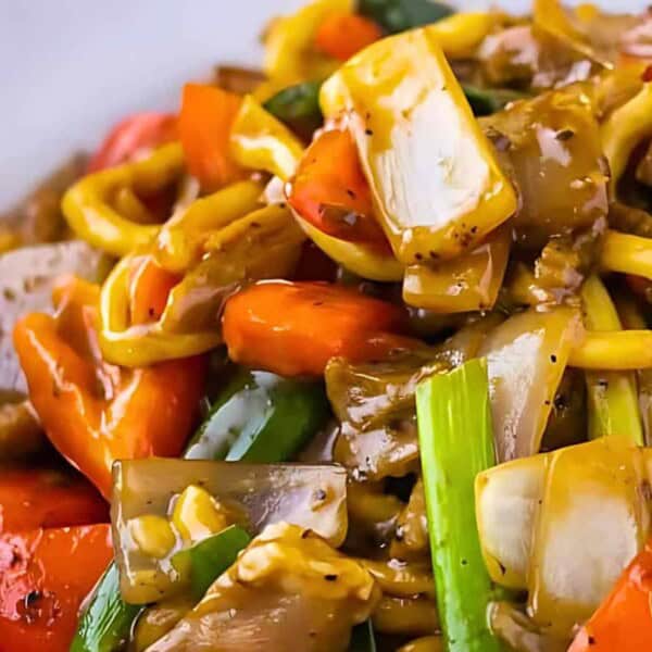 Close-up of a dish featuring stir-fried noodles, colorful bell peppers, onions, and other vegetables in a savory sauce on a white plate.