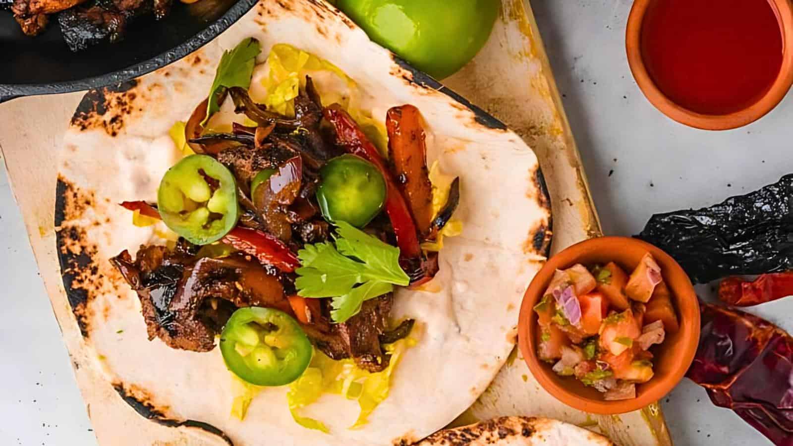 A tortilla topped with sliced grilled beef, bell peppers, jalapeños, and cilantro on a wooden board. Nearby are green tomatoes, a small dish of salsa, a cup of red sauce, and dried chili peppers.