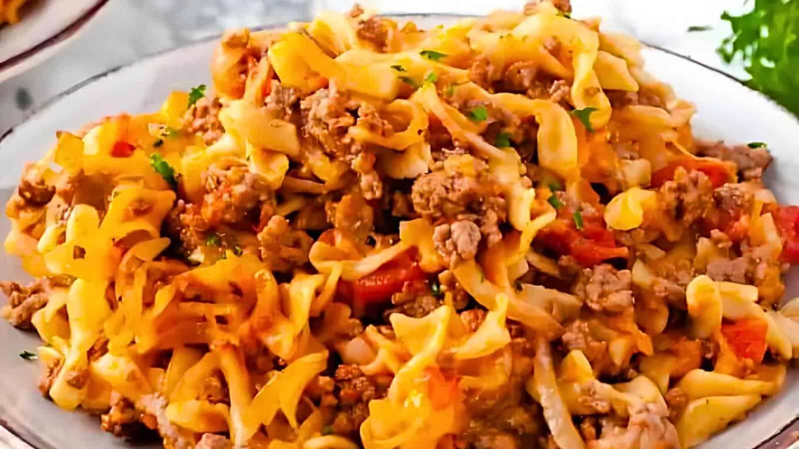 A plate of cheesy beef noodle casserole, featuring wide egg noodles mixed with ground beef, melted cheese, and chunks of red bell pepper, garnished with chopped parsley. The dish is served on a light-colored plate, with a blurred background.