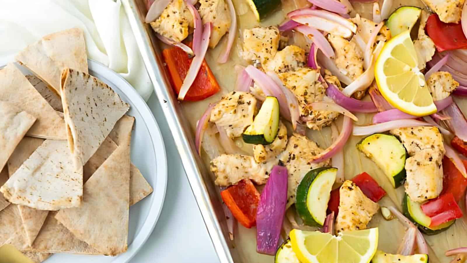 A sheet pan featuring baked chicken pieces with red and green bell peppers, red onions, and slices of lemon. The pan is accompanied by a plate of pita bread on the left side. The colorful, fresh ingredients create a vibrant and appetizing display.