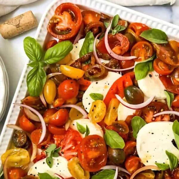 A rectangular plate of sliced tomatoes, mozzarella, red onions, basil, and oregano on a white surface. Four forks are placed beside the dish.