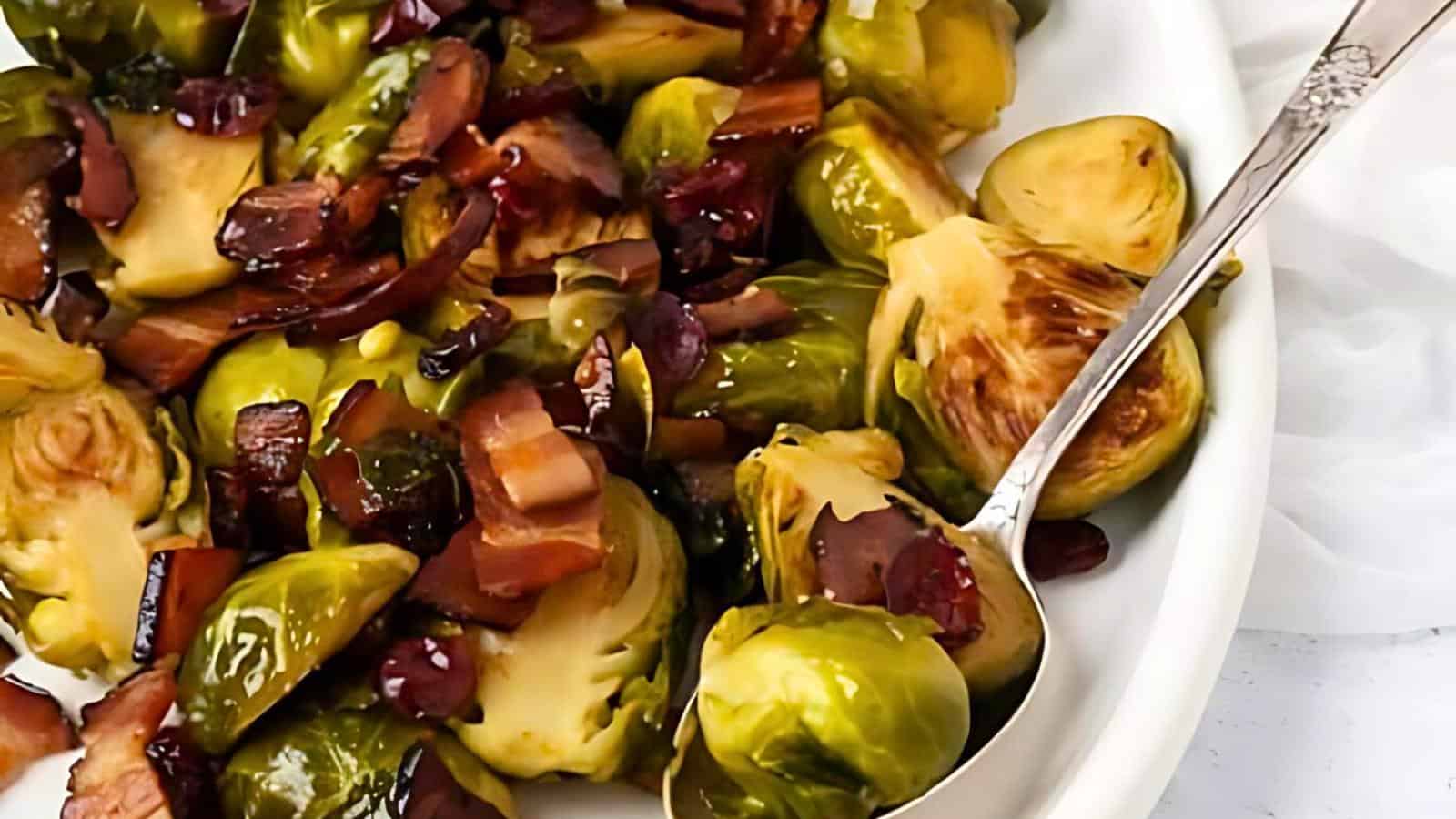 A white plate filled with roasted Brussels sprouts mixed with crispy bacon bits. A shiny silver spoon is placed on the right side of the plate, partially submerged in the mixture. The background includes a white surface and a partially visible white cloth.