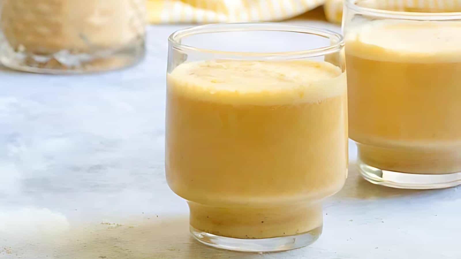 Two clear glasses filled with creamy yellow pineapple mousse are placed on a light-colored surface. Another container with mousse and a striped yellow cloth are visible in the background. The texture of the mousse looks smooth and inviting.
