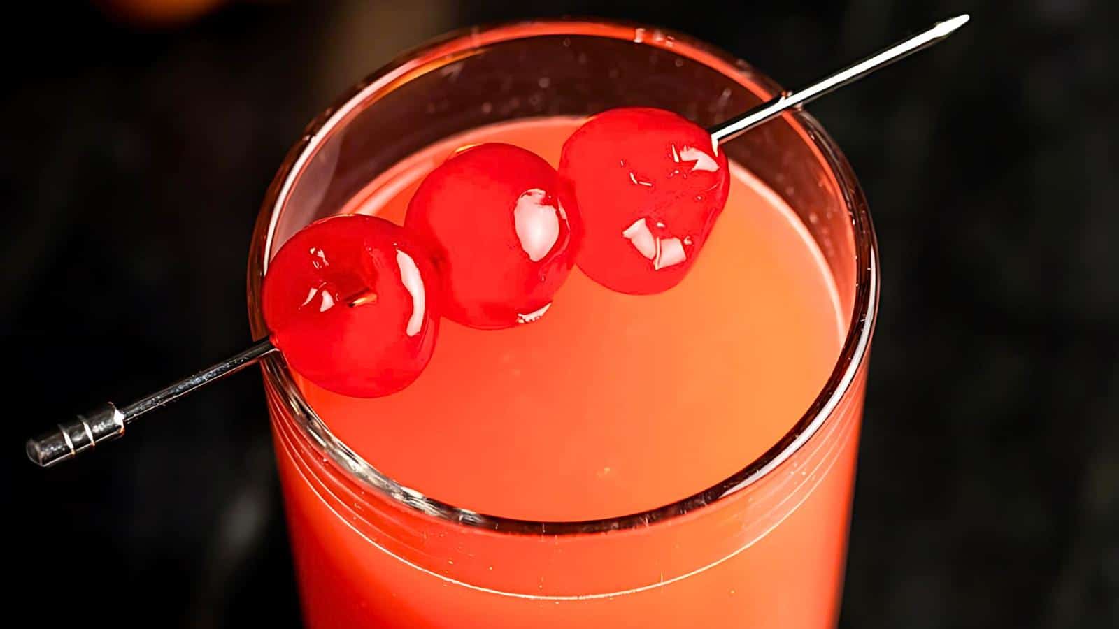 A tall glass filled with an orange-colored drink topped with three red cherries skewered on a metallic pick, placed horizontally across the rim of the glass. The background is dark, which accentuates the vibrant colors of the drink and cherries.