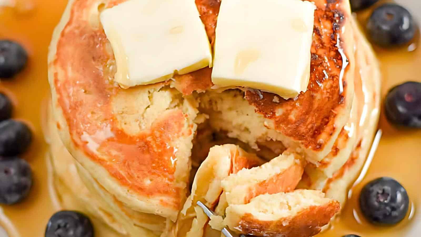 A close-up of a stack of fluffy pancakes topped with two pats of butter. A fork is lifting a bite from the stack, revealing the tender interior. Blueberries are scattered around the pancakes, and the dish is generously drizzled with syrup.