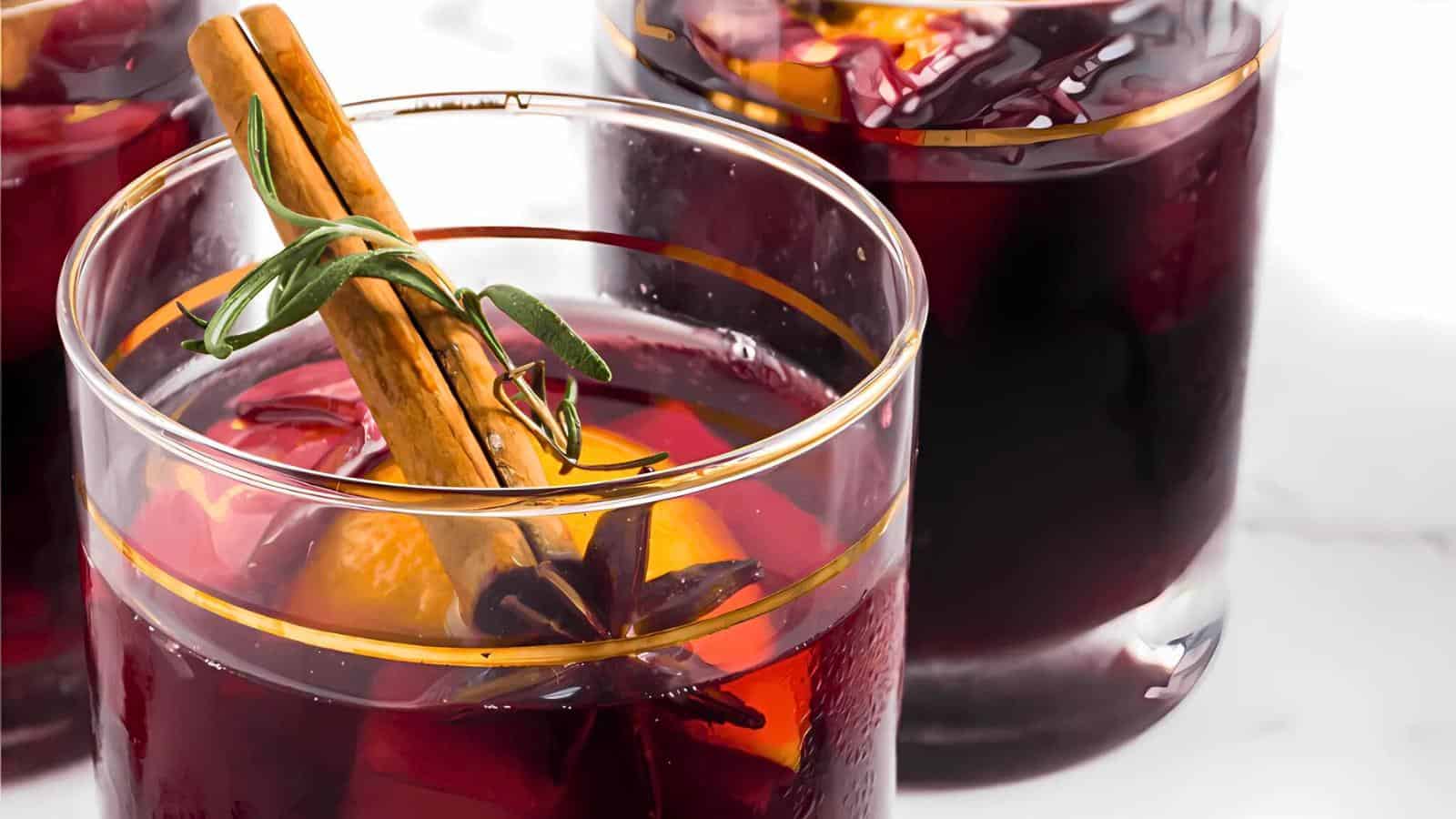 Close-up of glasses filled with mulled wine garnished with cinnamon sticks, orange slices, star anise, and fresh herbs. The red wine appears rich and inviting, with aromatic spices enhancing its presentation. The background is softly blurred, emphasizing the drinks.