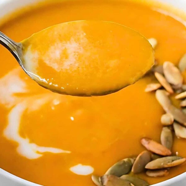 A bowl of creamy harvest soup topped with pumpkin seeds and a swirl of cream, with a spoonful held above it.