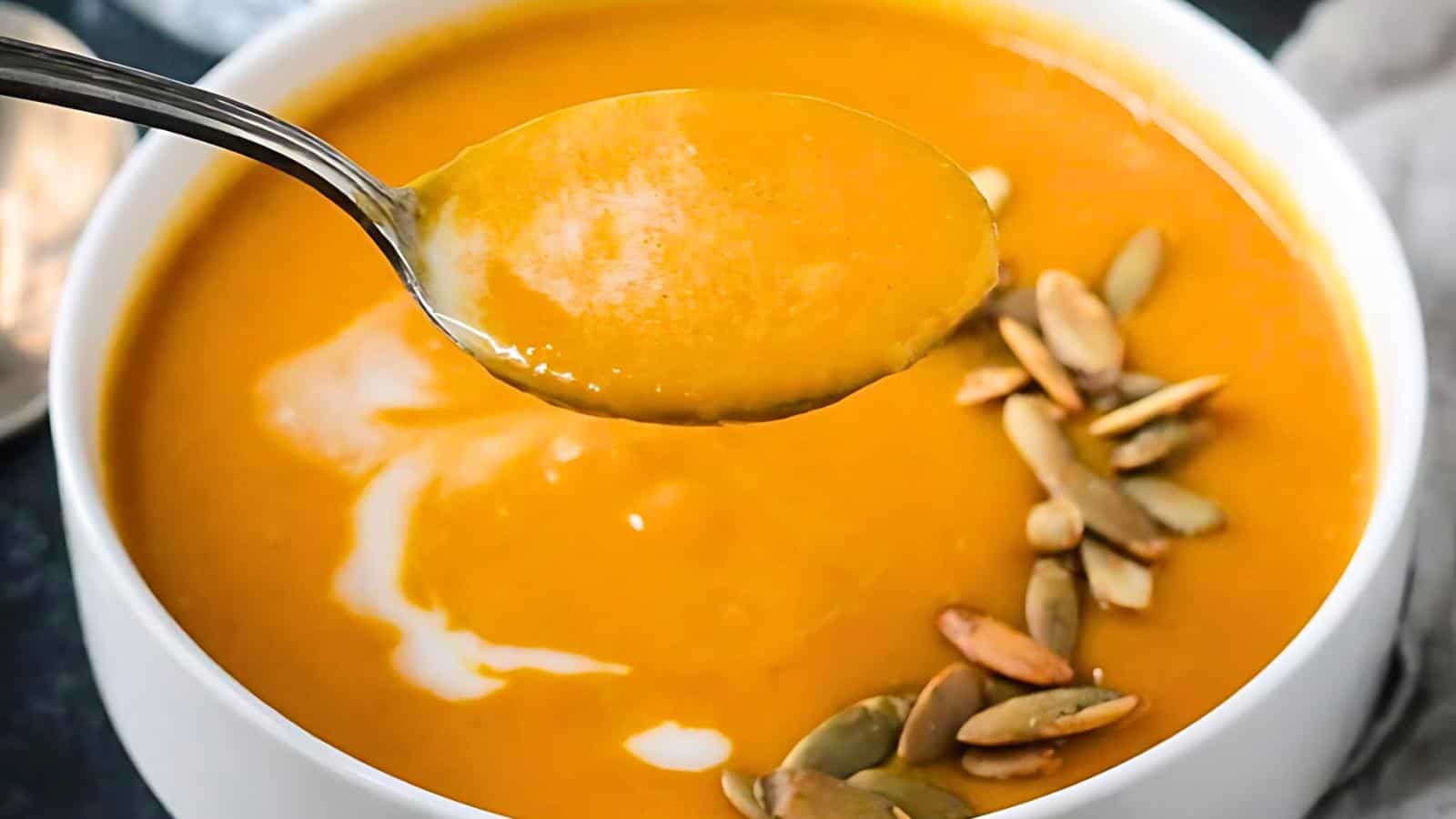 A spoonful of creamy orange-colored soup is held above a bowl filled with the same soup. The bowl is garnished with a drizzle of white cream and topped with a cluster of green pumpkin seeds on the surface.